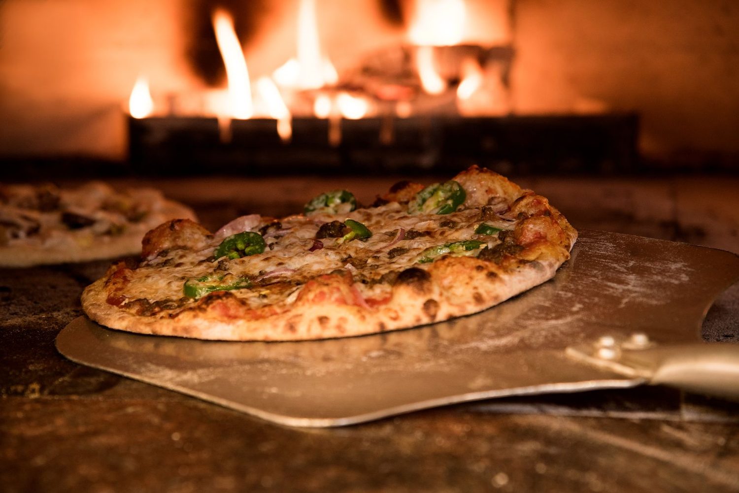 Pizza artesanal saindo do forno a lenha com queijo derretido e chama ao fundo